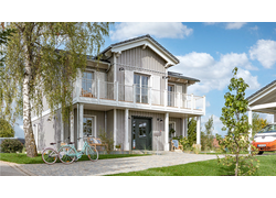 Frontansicht des Hauses. Die komplette Vorderseite ist überdacht durch einen Balkon, die Fassade ist aus Holz. Rechts sieht man den Carport. 