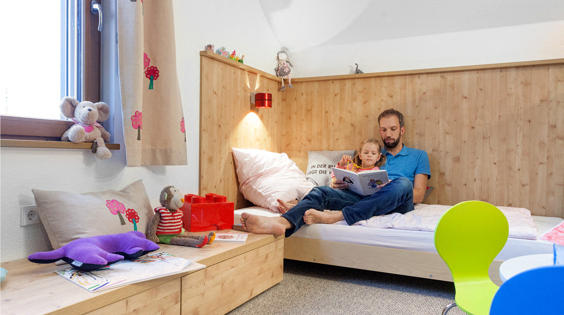 Helles Kinderzimmer mit großem Bett auf welchem der Papa seiner Tochter etwas vorliest.